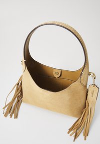 Sac à main en suede tan, avec une poignée courbée, une poche intérieure, des accents de pompon et une fermeture à pression. Présente des détails en métal doré.