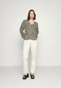 Blusa verde a trama con scollo a V, chiusura con bottoni, maniche lunghe, abbinata a pantaloni bianchi e tacchi neri spessi.