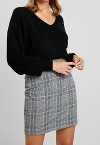 Schwarzer kabelgestrickter Pullover mit V-Ausschnitt, kombiniert mit einem karierten Rock in Grau-, Blau- und Schwarztönen. Enganliegende Silhouette.