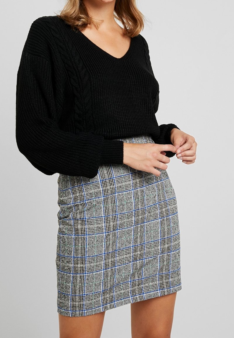 Schwarzer kabelgestrickter Pullover mit V-Ausschnitt, kombiniert mit einem karierten Rock in Grau-, Blau- und Schwarztönen. Enganliegende Silhouette.