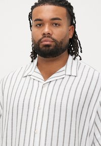 Λευκό λινό πουκάμισο με λεπτές γκρι κάθετες ρίγες, κοντό γιακά και κλείσιμο με κουμπιά μπροστά. Ελαφριά υφή, casual σχεδιασμός.