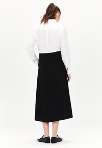 Witte overhemd met lange mouwen en knoopmanchetten; zwarte A-lijn rok die tot halverwege de kuit reikt; model draagt zwarte instappers; effen achtergrond.