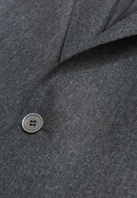 Tessuto di lana grigia con una texture liscia, caratterizzato da un unico bottone lucido con quattro fori. Primo piano del bavero e della tasca della giacca.