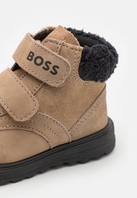Brune suède ankelstøvler med sort fleeceforing, velcrorem og struktureret sort gummisål. Mærkenavnet "BOSS" vises på remmen.