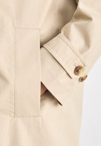 Beige trench coat with a smooth texture, featuring a rectangular patch pocket and two round, patterned buttons on the cuff.