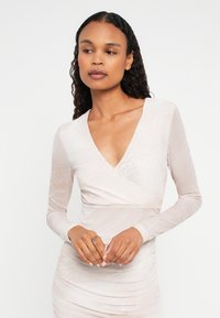 Γυναίκα με μακριά κυμματιστά μαλλιά, φορώντας ένα ελαφρύ κρεμ μανίκι φόρεμα με ρίχτι και διακριτικό ζώον σχέδιο, κοιτάζοντας ελαφρώς προς το πλάι με τα χέρια ενωμένα.