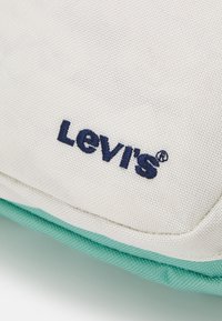 Levi's toteväska har en beige canvasutsida med en teal plastdetalj och ett marinblått broderat logotyp. Strukturerad tyg med sömnadsdetaljer.