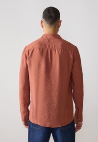 Chemise à manches longues couleur rouille avec une coupe décontractée, dotée d'un col à boutons et de manches complètes. Texture lisse avec un léger éclat.