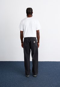 Schwarze Jeans mit Nadelstreifen und entspannter Passform, kombiniert mit einem schlichten weißen T-Shirt mit kurzen Ärmeln. Sichtbares Markenpatch auf der hinteren Tasche.