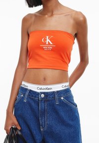 Orange strapless crop top med vitt "ck"-logotyp, matchad med högmidjade blå denimjeans som har en vit Calvin Klein-linning.
