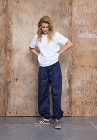 Kvinde står og kigger ned med hænderne på hofterne, iført hvid t-shirt, mørkeblå bukser og brune sneakers mod en træbaggrund.