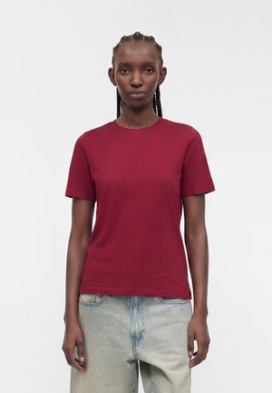 Femme aux cheveux tressés portant un t-shirt bordeaux uni à manches courtes et un jean bleu clair sur un fond neutre.