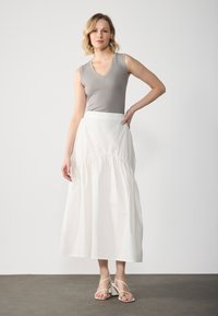 Haut sans manches gris associé à une jupe longue blanchi, froncée. Le mannequin porte des sandales à talons de couleur claire. Design simple et épuré sur un fond neutre.