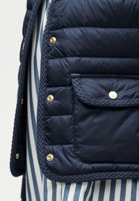 Τσέπη ναυτικού μπουφάν με καπιτονέ, πλεκτές λεπτομέρειες και χρυσά κουμπιά με τρούκ, πάνω από ύφασμα με μπλε και λευκές ρίγες.
