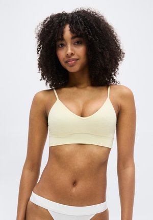 Kobieta o kręconych włosach, ubrana w jasnobeżowy biustonosz typu bralet i białą bieliznę, stojąca na jednolitym białym tle.