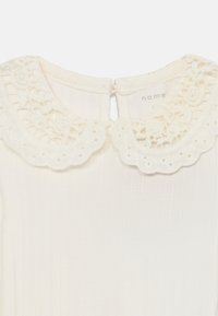 Robe crème avec un tissu texturé, présentant un collier en dentelle à frange et une fermeture à boutons à l'arrière.