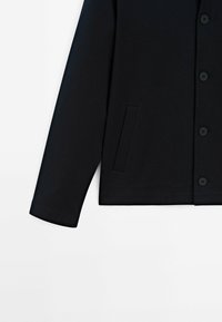 Giacca nera con una texture liscia, dotata di una chiusura a bottoni, una tasca laterale e maniche a taglio dritto.
