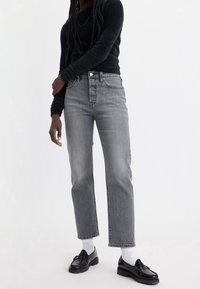 Persona con un suéter negro de peluche, jeans grises de cintura alta, calcetines blancos y mocasines negros, de pie contra un fondo blanco.