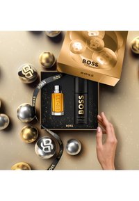 Scatola nera contenente una bottiglia di colonia oro e nera e uno spray deodorante, circondata da sfere dorate e argentate, con un nastro.