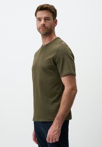 T-shirt verde oliva a maniche corte con collo rotondo, forma aderente e texture morbida simile al cotone, indossato da un uomo di lato.