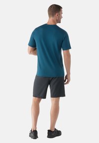 Camiseta deportiva de manga corta en color verde azulado con cuello redondo, combinada con pantalones cortos negros y zapatillas negras, vista desde la parte posterior. Textura suave.