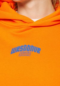 Gros plan d'une personne portant un sweat à capuche orange vif avec le texte bleu "westblue studio" sur la poitrine, montrant le cou et une partie de la mâchoire.