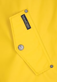 Jasnożółta tkanina z klapkową kieszenią zapinaną na trzy metalowe zatrzaski oraz czarną metką z napisem "Schmuddelwedda".