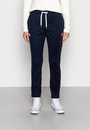 Persoon draagt marineblauwe smalle sweatpants met witte trekkoord, gecombineerd met witte hoge sneakers en een witte geribde trui.