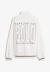 Bela kapuca s visokim ovratnikom, s podebljanim crnim tekstom koji glasi "NAPRAVLJENO ZA HRABRE I NEUSTRAŠIVE." Rebrasti smeđi detalji na manžetama.