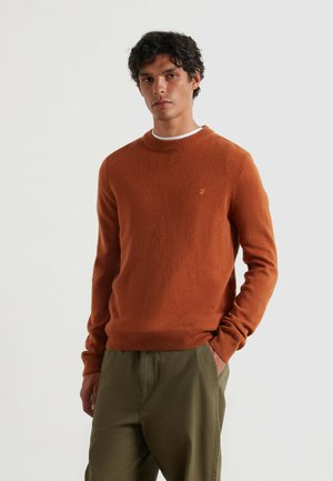 Pull en maille orange rouille avec un col rond et des poignets côtelés. Doté d'un petit logo brodé sur la poitrine. Associé à un pantalon vert ample.