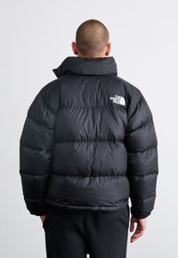 Crna puf jakna s visokim ovratnikom, s prošivenim dizajnom i elastičnim rukavima. Logo The North Face prikazan je u bijeloj boji na lijevom ramenu.