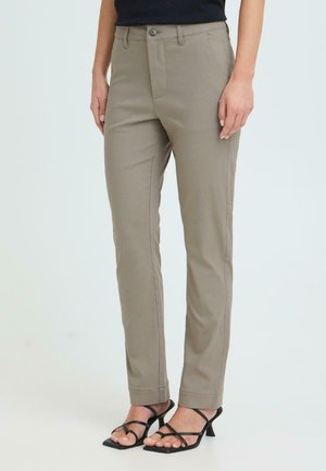 Person iført beige bukser med rett ben og svarte sandaler med hæl og stropper, stående mot en enkel, lys bakgrunn.