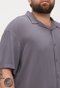 Camisa de botones de manga corta en gris, con cuello clásico y tejido suave. Presenta botones negros y un corte relajado.