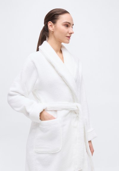 Νέα γυναίκα με καστανοκόκκινα μαλλιά σε αλογοουρά, φορώντας λευκή αφράτη ρόμπα με γιακά και ζώνη στη μέση, με το χέρι στην τσέπη, κοιτάζοντας προς τα δεξιά.