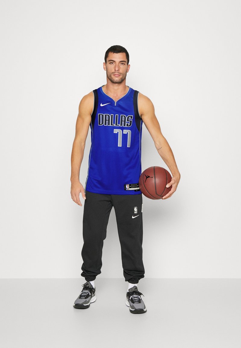 Niebieski strój koszykarski z napisem "DALLAS" i numerem "77" w białym kolorze. Czarne spodnie dresowe, szare sneakersy, trzymający piłkę do koszykówki. Prosty, sportowy design.