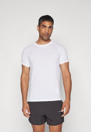 Camiseta deportiva blanca de manga corta, con cuello redondo y un logotipo sutil. Textura suave; combinada con pantalones cortos deportivos oscuros.