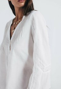 Hvid linned bluse med lodrette plissee, V-udskæring og blonderammelser på ærmerne. Blød tekstur med svævende designelementer.