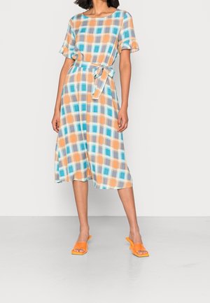 Femme portant une robe mi-longue à carreaux orange et bleus avec ceinture et des talons ouverts orange, debout devant un fond blanc.