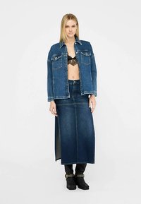 Chaqueta vaquera con dos bolsillos en el pecho, combinada con una falda larga de mezclilla con una abertura lateral. Color azul oscuro, combinada con botas negras.