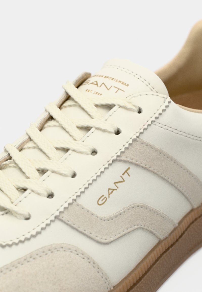 Weiße Sneaker mit Obermaterial aus Wildleder und Leder, beigen Akzenten, strukturierten Schnürsenkeln und goldfarbenem "GANT"-Logo an der Seite. Gummisohle sichtbar.