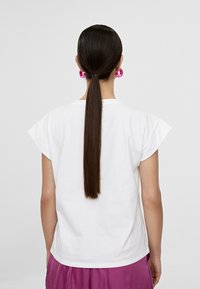 Vit t-shirt med kort ärm och uppvikta ärmar, kombinerad med rosa byxor. Långt rakt hår som är säkrat med ett hårband.