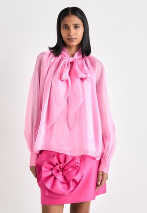 Rosa, gjennomsiktig bluse med en stor sløyfe ved halsen og lange ermer, kombinert med en rosa skjørt med et 3D-blomsterdesign ved hoften.