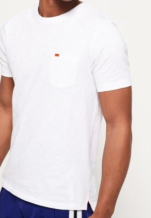 Mężczyzna ubrany w zwykły biały krótki rękaw z kieszonką t-shirt oraz niebieskie spodnie z białymi paskami po bokach, stojący na białym tle.