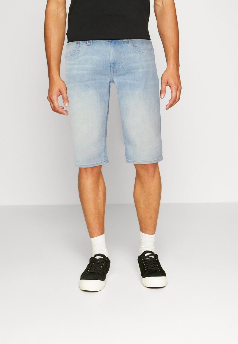 Shorts de mezclilla azul claro con un ajuste relajado, que llegan justo por encima de las rodillas, combinados con calcetines tobilleros blancos y zapatillas negras.