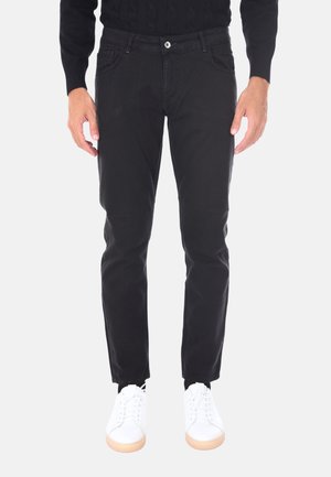 Uomo in piedi che indossa pantaloni neri, sneakers bianche e una maglia nera a maniche lunghe, mostrato dalla vita in giù su uno sfondo bianco.
