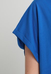 Detail rukávu modrého oděvu zobrazující texturu látky a paži osoby na jednoduchém světle šedém pozadí.