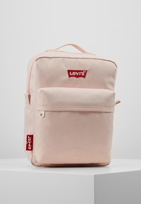 Vaalea vaaleanpunainen kangaskäyttöön suunniteltu reppu, jossa on etutasku, punaiset Levi's-logo yksityiskohdat ja vetoketju, suorakulmainen muoto ja tukevat kahvat.