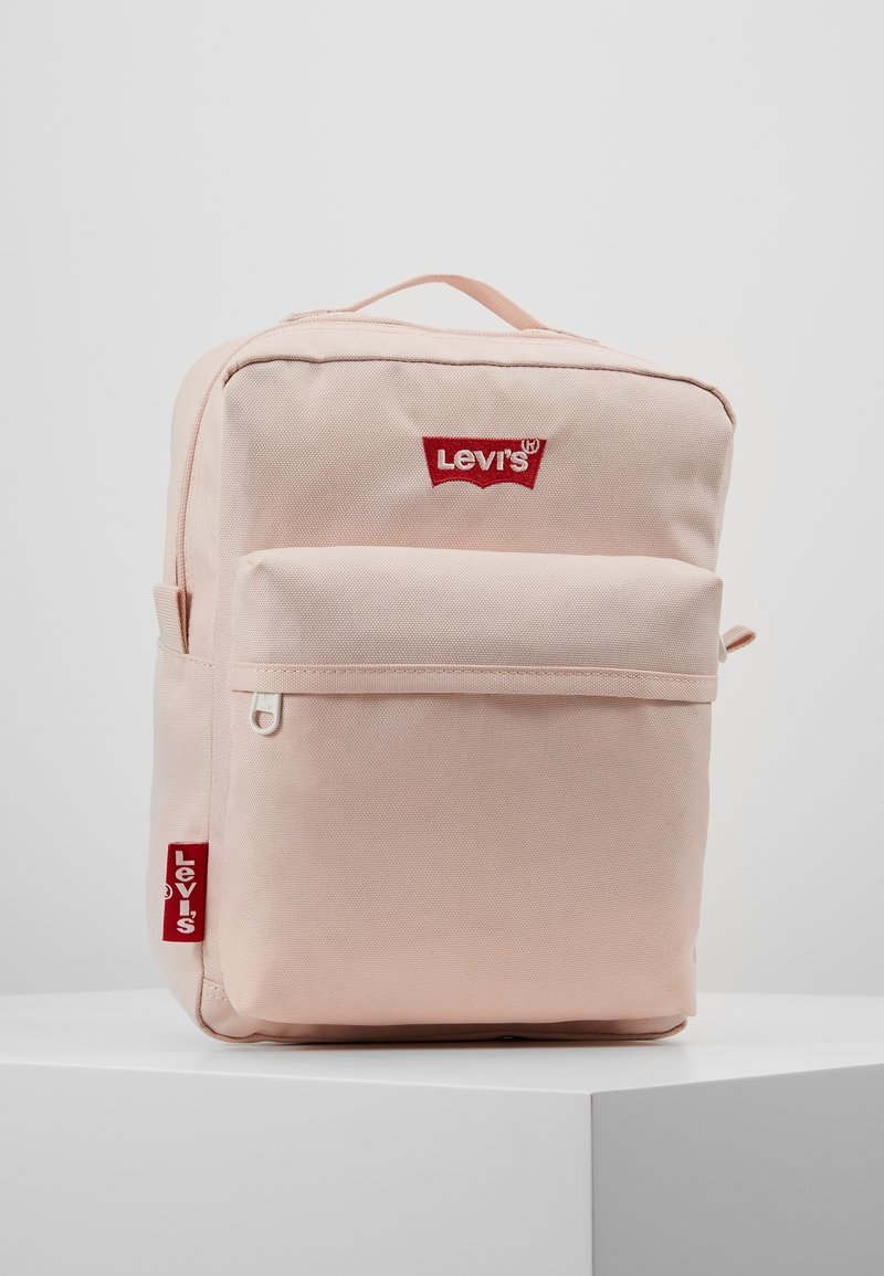 Vaalea vaaleanpunainen kangaskäyttöön suunniteltu reppu, jossa on etutasku, punaiset Levi's-logo yksityiskohdat ja vetoketju, suorakulmainen muoto ja tukevat kahvat.