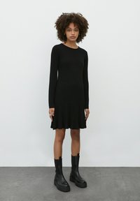 Schwarzes, geripptes Langarmkleid mit schwingendem Saum. Der Stoff hat eine strukturierte Textur. Kombiniert mit schwarzen Stiefeln mit dicker Absatzhöhe. Einfacher runder Ausschnitt.