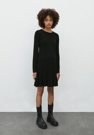Robe noire à manches longues et côtelées avec un ourlet évasé. Le tissu est texturé. Associée à des bottes noires à talons épais. Encolure ronde simple.
